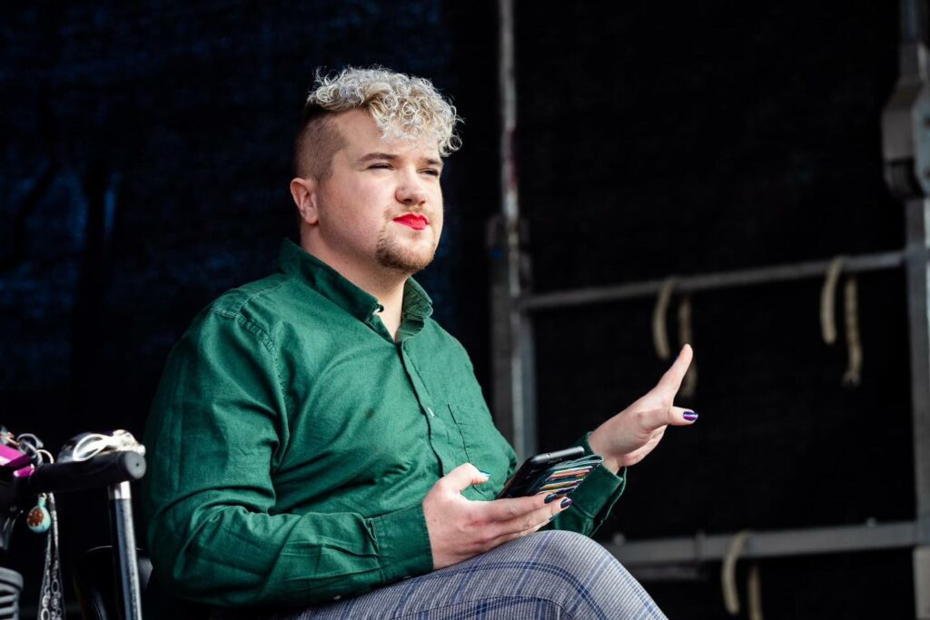 A person with make-up on sits on a stage, holding a phone and their hand out right.