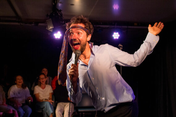 A man with his tie tied to his head smiling with his arms open.