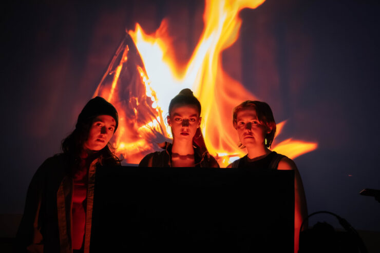 Three young people stand behind a fire projection looking towards the camera.
