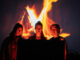 Three young people stand behind a fire projection looking towards the camera.