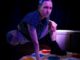 a trans person with a blue patterned buzz cut plays a game of twister. their left knee bent and right hand stabalising it