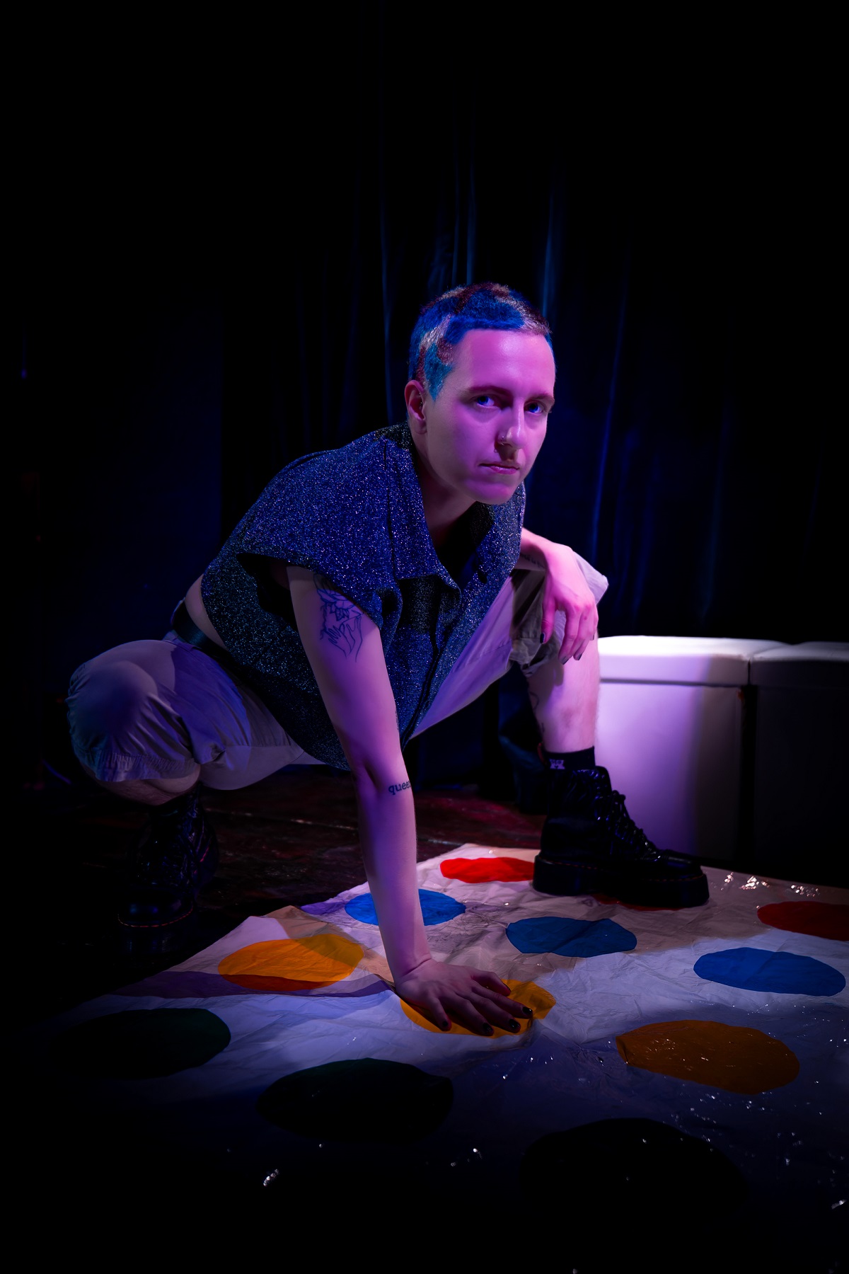 a trans person with a blue patterned buzz cut plays a game of twister. their left knee bent and right hand stabalising it