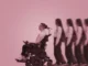A non-binary person with a brown buzz cut is sat in an electric wheelchair going towards the left of the image, behind them is a woman with long curly hair wearing a backpack following them, she gets more blurry towards the edge of the image