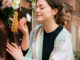a woman with mid length brown hair in a ponytail, wearing a black t shirt and floral kimono stands outside an old brick house. Closing her eyes as she smells the flowers draping over it, holding her electric guitar against the wall.