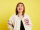 An Asian woman with mid length ginger hair wearing a white jersey jacket and a black t shirt smiling looking just off the camera. She is standing in front of a bright yellow background