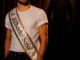Jilberto Soto, male with short blonde hair and a brown beard wears an ornate crown and sash reading 'Mr Jilberto Soto' is smiling and looks just off the camera. behind him is a spotlighted silhouette of a person in a sombrero