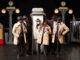 Five black men are standing at a 1920s telephone booth. all of them in the similar outfits, the three at the front wear black trilby hats, beige half trench coats, blue blazers with a matching tie over a white shirt tucked into black trousers and smart black shoes but the two are the back wear brown leather bomber jackets with the same black trilby hats, blue ties and black sunglasses. There is one of the brothers in the middle on the phone laughing, holding a rose. The others are looking at him with neutral expressions