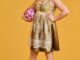 A trans woman with mid length curly brown hair stands in the centre of a yellow background in a gold dress holding a white and ink football in one hand wearing white and black stripped football socks and bright pink football boots. She stands in determination looking just off the camera
