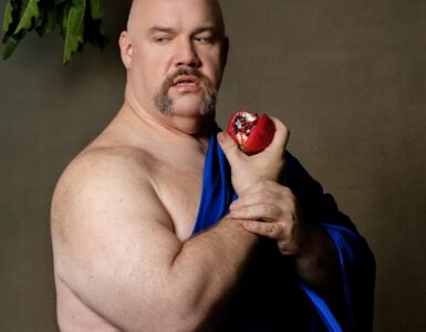 A plus size bald man wears a bright dark blue fabric over his chest, he holds a bitten pomegranate. He looks just off the camera