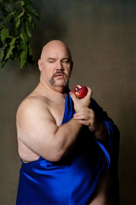 A plus size bald man wears a bright dark blue fabric over his chest, he holds a bitten pomegranate. He looks just off the camera