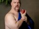 A plus size bald man wears a bright dark blue fabric over his chest, he holds a bitten pomegranate. He looks just off the camera