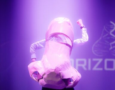 A man stands in a paper mache nose costume over the top of a white shirt and black trousers, his left arm at a 90 degree angle pointing to the floor and his right arm at a 90 degree angle pointing upwards