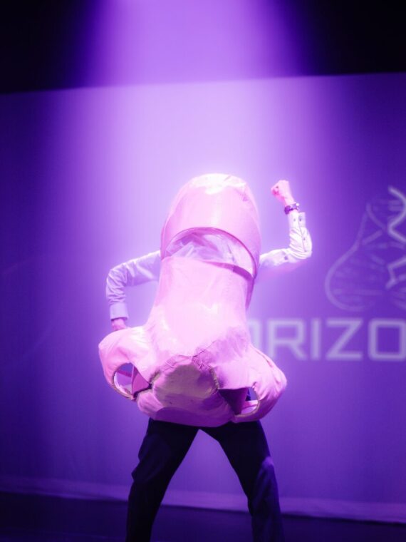 A man stands in a paper mache nose costume over the top of a white shirt and black trousers, his left arm at a 90 degree angle pointing to the floor and his right arm at a 90 degree angle pointing upwards
