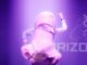 A man stands in a paper mache nose costume over the top of a white shirt and black trousers, his left arm at a 90 degree angle pointing to the floor and his right arm at a 90 degree angle pointing upwards