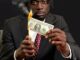 A black man with short locs sits at a smart varnished table wearing a black suit jacket, white shirt and red patterned tie. He holds a lighter as he burns the edge of a dollar note staring at it in concentration as he does so