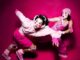 two feminine presenting people are pulling each other away from something coming from the left. Natasha (left) has black curly hair with bright white face paint with bright pink rosy cheeks. She's wearing a light pink jumper, bright pink fingerless gloves, bright pink shorts with light pink knee pads and bright pink lace up sneakers. She is being pulled by Xhloe who also has black curly hair with the same face paint but wears a light pink beanie, bright pink overalls, elbow pads and light pink lace up shoes. they both look to the left, scared
