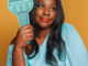 A black woman with black hair poses in front of a yellow background wearing a blue dress and crown holding the statue of Liberty's torch