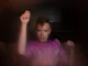 Aidan, a man with short brown hair saved at the sides with a few curls hanging over his face is wearing a pink velvet jumper looking directly into the camera, his left arm up towards the ceiling just about to click.
