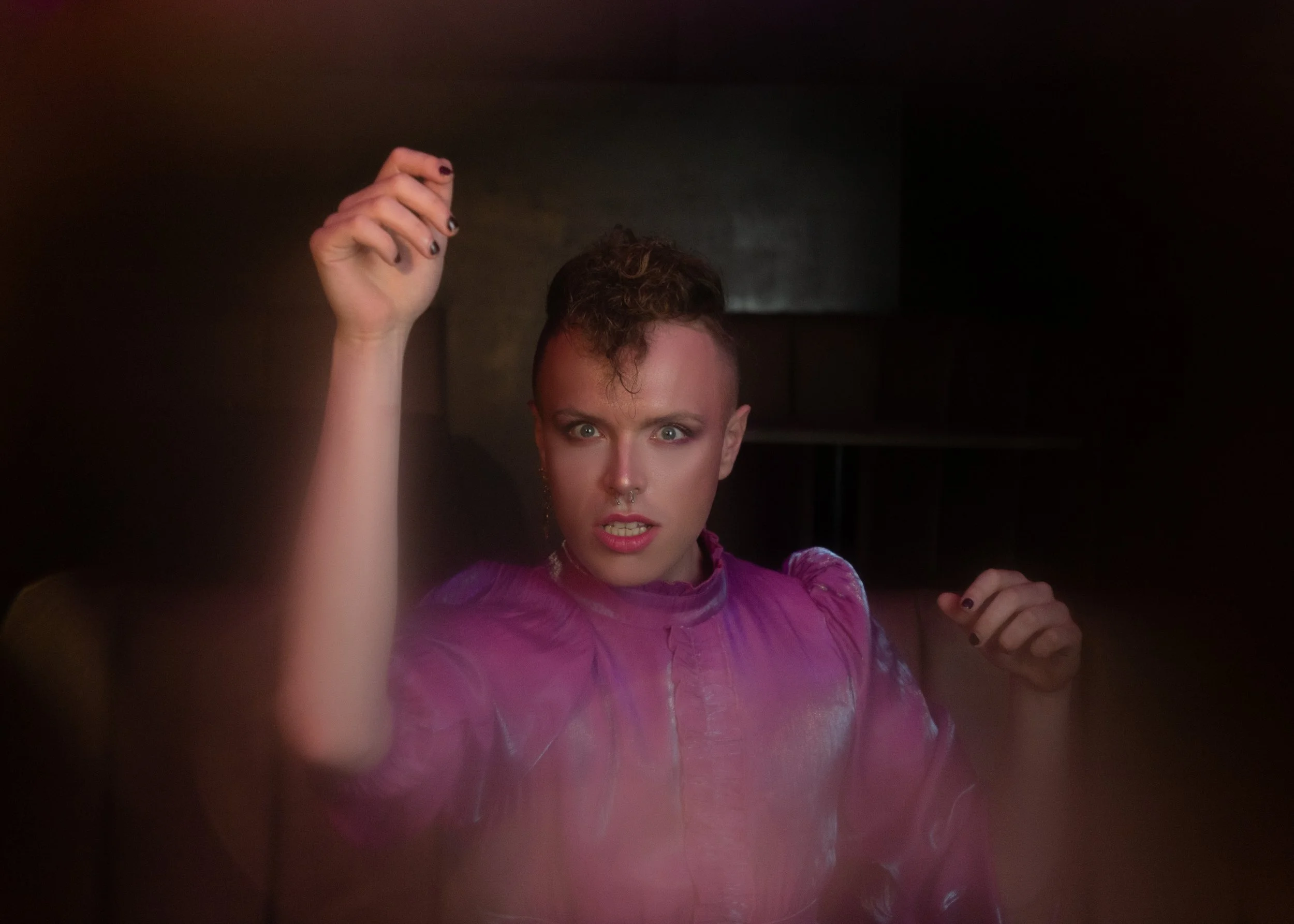 Aidan, a man with short brown hair saved at the sides with a few curls hanging over his face is wearing a pink velvet jumper looking directly into the camera, his left arm up towards the ceiling just about to click.