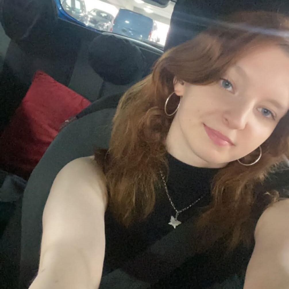 a woman sits in the front seat of a car. she has mid length wavy brown hair, silver hoop earrings and a sleeveless black top. She wears a silver butterfly necklace and her arms reach up to hold the phone the photo is taken on