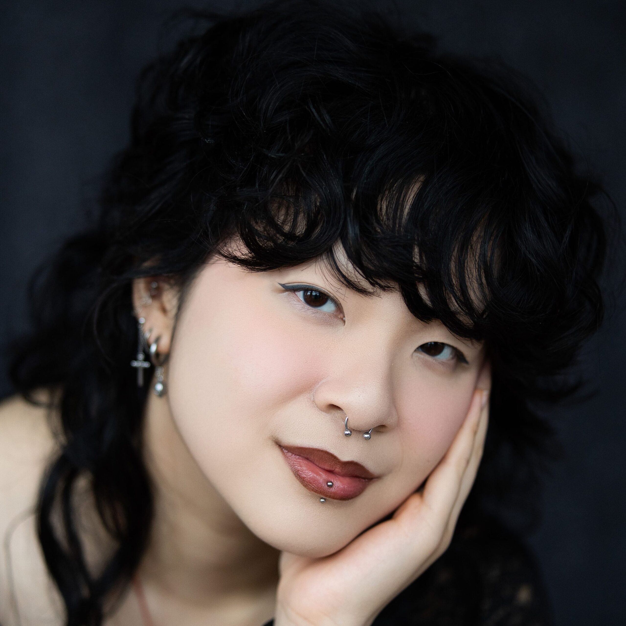 A headshot of a non-binary person with a black straight long mullet. They have multiple earrings in their left ear. Their head is leaning into their right hand as they smile at the camera