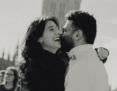 A black and white photo of a man and a woman hugging. She is laughing, and in the background you can see Big Ben.