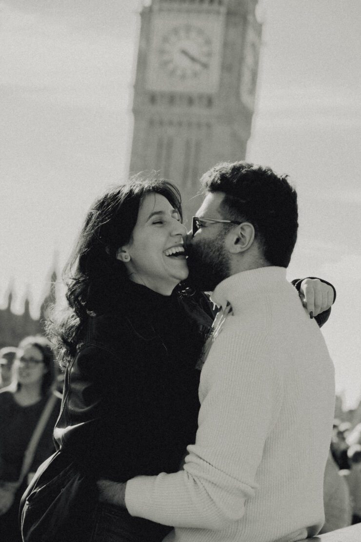 A black and white photo of a man and a woman hugging. She is laughing, and in the background you can see Big Ben.