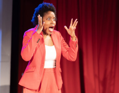 A woman wearing a red suit jacket and pants holds her hands up near her face with a shocked look.