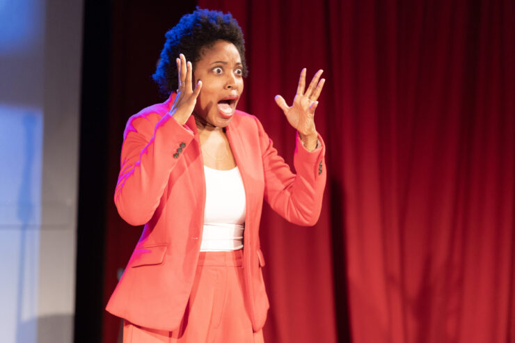A woman wearing a red suit jacket and pants holds her hands up near her face with a shocked look.
