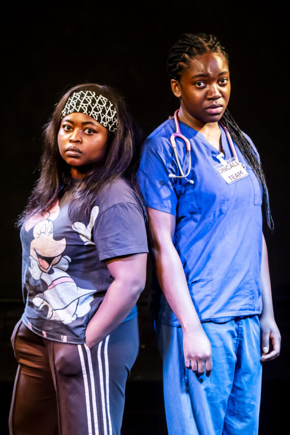 Two women face the camera their backs to each other. Woman on the left is wearing casual clothing and woman on the right wearing scrubs.