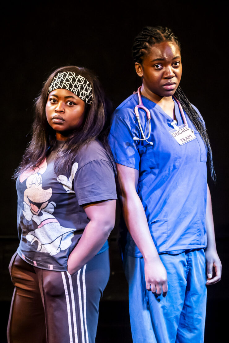 Two women face the camera their backs to each other. Woman on the left is wearing casual clothing and woman on the right wearing scrubs.