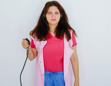 A woman swearing a pink suit, blue jeans and a pink spotted sleeveless smock holds a blood pressure bulb in her right arm.