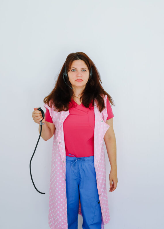 A woman swearing a pink suit, blue jeans and a pink spotted sleeveless smock holds a blood pressure bulb in her right arm.