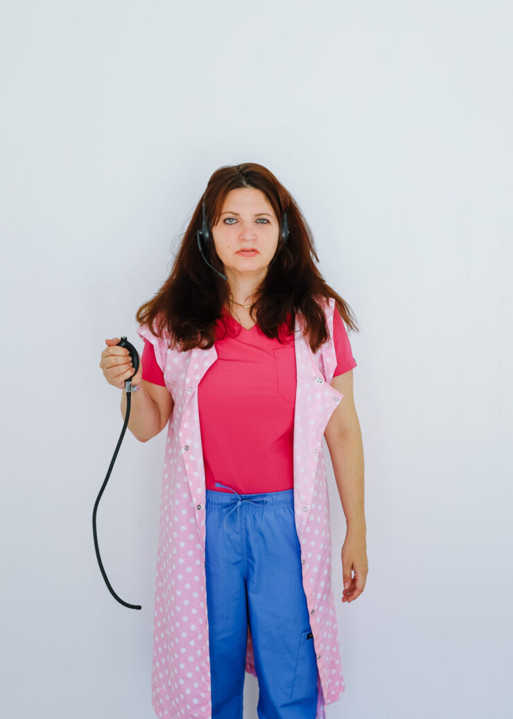A woman swearing a pink suit, blue jeans and a pink spotted sleeveless smock holds a blood pressure bulb in her right arm.