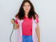 A woman swearing a pink suit, blue jeans and a pink spotted sleeveless smock holds a blood pressure bulb in her right arm.