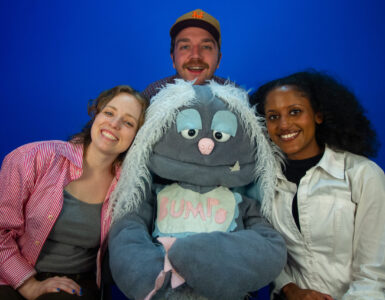 A grey puppet with light blue hair is surrounded by 3 people, on their left, right and directly behind the puppet.
