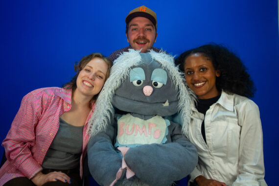 A grey puppet with light blue hair is surrounded by 3 people, on their left, right and directly behind the puppet.