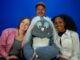 A grey puppet with light blue hair is surrounded by 3 people, on their left, right and directly behind the puppet.