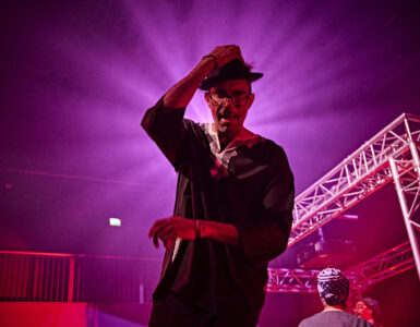 A man is touching his hat on his head with his right hand. Behind his is a bring pink glowing source illuminating the image.