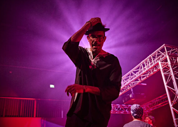 A man is touching his hat on his head with his right hand. Behind his is a bring pink glowing source illuminating the image.