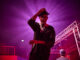 A man is touching his hat on his head with his right hand. Behind his is a bring pink glowing source illuminating the image.