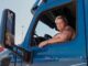 a woman with a tattoo and short blonde hair leans out of the window of a big-rig truck cabin. Terri Solem by Anne-Marie Michell