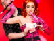 A woman wearing a pink corset, holds a magicians cane in a young man's mouth as he holds playing cards behind a red background.