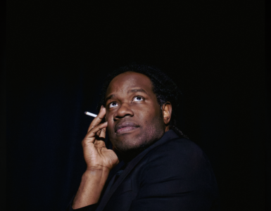a young Black man holds his right cheek with a cigarette between those fingers looking off to the right side of the image wearing black and a black background.