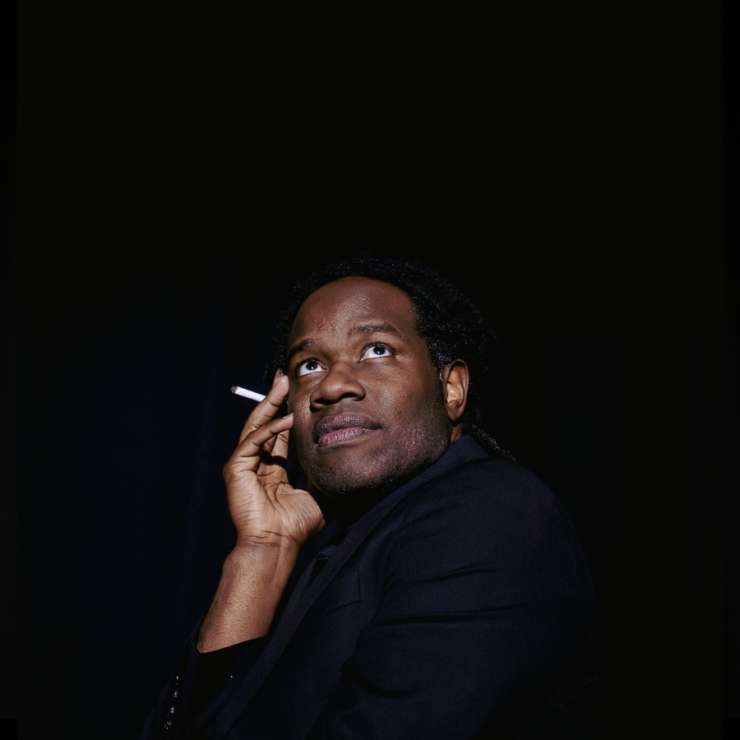 a young Black man holds his right cheek with a cigarette between those fingers looking off to the right side of the image wearing black and a black background.