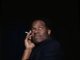 a young Black man holds his right cheek with a cigarette between those fingers looking off to the right side of the image wearing black and a black background.